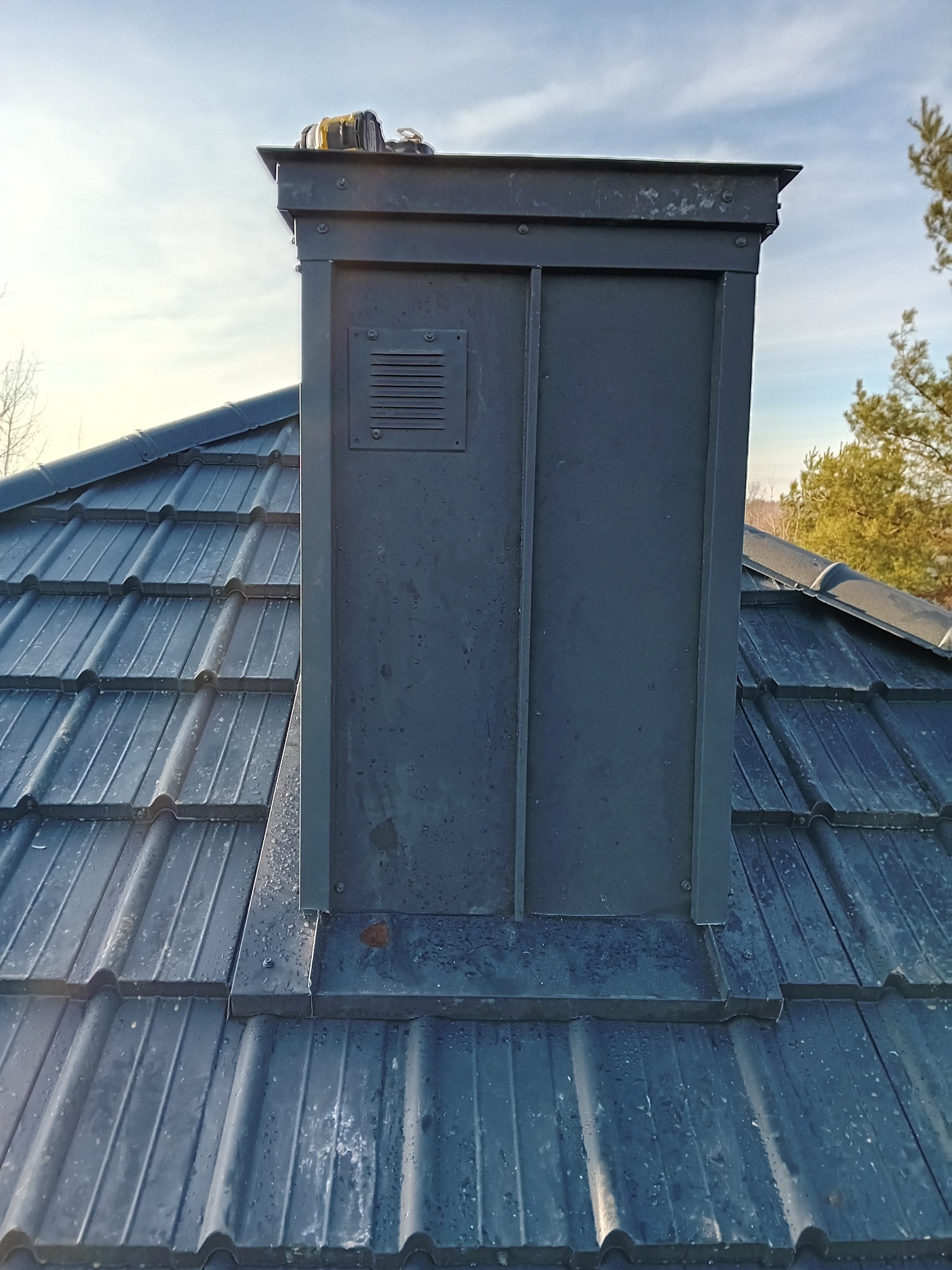 Metalowy komin z kratką wentylacyjną na dachu z ciemnej dachówki ceramicznej, widok z dołu, narzędzia na górze komina, drzewa w tle.