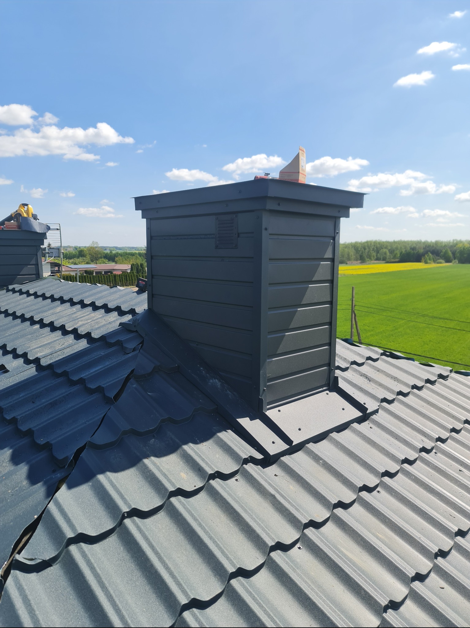 Szary komin wentylacyjny na dachu z blachodachówki, na tle pól i błękitnego nieba. Usługi dekarskie w Tczowie.