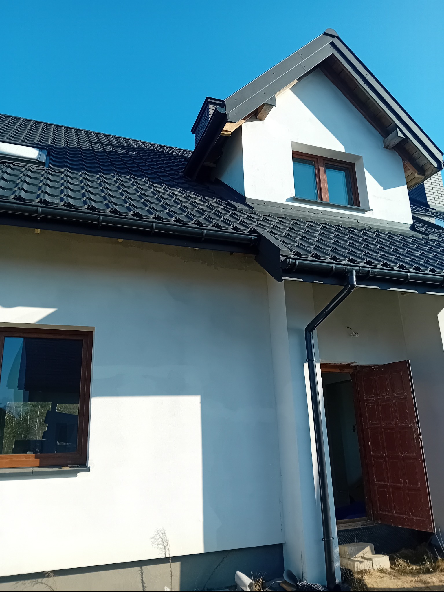 Dom jednorodzinny z nowym, czarnym dachem z dachówki ceramicznej i zamontowanymi rynnami. Widok na fragment elewacji i otwarte drzwi wejściowe.