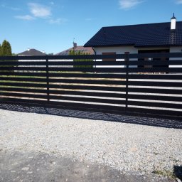 KRYMART Kowalstwo Artystyczne Krzysztof Misiewicz - Nowoczesna, przesuwna brama w kolorze antracytu z poziomymi panelami, na tle domu z ciemnym dachem i błękitnego nieba. Podłoże żwirowe.