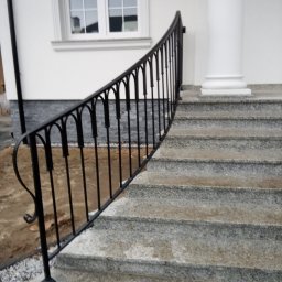 KRYMART Kowalstwo Artystyczne Krzysztof Misiewicz - Elegancka, czarna, metalowa balustrada schodowa z ozdobnymi łukami, zamontowana przy granitowych stopniach wejściowych do budynku z kolumną.
