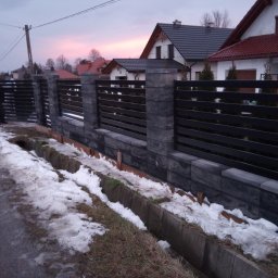 KRYMART Kowalstwo Artystyczne Krzysztof Misiewicz - Nowoczesne ogrodzenie panelowe z elementami betonowymi w odcieniach szarości, widoczne na tle domów jednorodzinnych o zmierzchu. Ślady śniegu wzdłuż ogrodzenia.