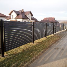 KRYMART Kowalstwo Artystyczne Krzysztof Misiewicz - Nowoczesne, poziome ogrodzenie panelowe w kolorze antracytowym, oddzielające posesję z domem o brązowym dachu. Widok z perspektywy ścieżki.