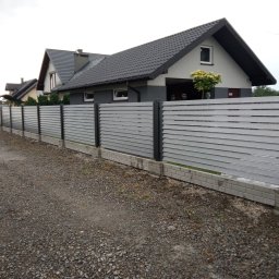 KRYMART Kowalstwo Artystyczne Krzysztof Misiewicz - Nowoczesne ogrodzenie panelowe w kolorze szarym, z podmurówką z jasnej cegły, na tle domu z ciemnym dachem. Widok z perspektywy żwirowej drogi.
