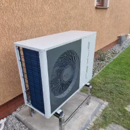 Biała pompa ciepła marki NEXLUS EVI, zamontowana na metalowym stelażu na betonowej podstawie, na tle otynkowanej ściany budynku i trawnika.