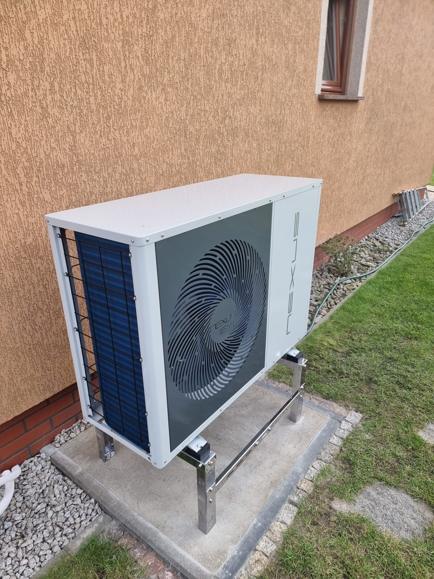 Biała pompa ciepła marki NEXLUS EVI, zamontowana na metalowym stelażu na betonowej podstawie, na tle otynkowanej ściany budynku i trawnika.
