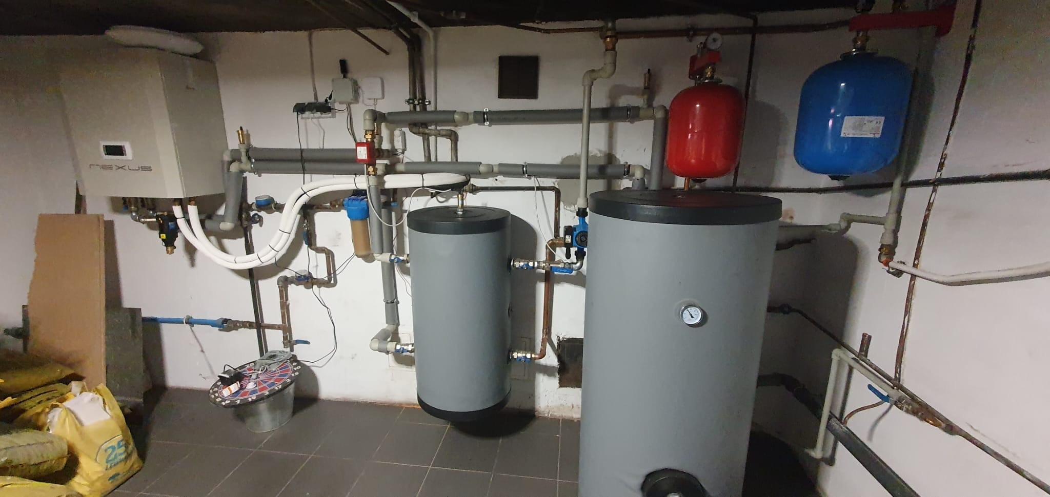 Instalacja w piwnicy z dwoma szarymi zbiornikami akumulacyjnymi, czerwonym i niebieskim naczyniem wzbiorczym, kotłem gazowym Nexus, oraz licznymi rurami i zaworami.