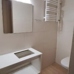 Jasna łazienka z beżowymi płytkami, białą szafką pod umywalkę z blatem z otworem na umywalkę, grzejnikiem drabinkowym i lustrem z oświetleniem.