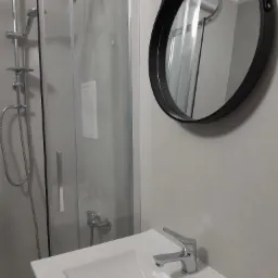 Nowoczesna łazienka z kabiną prysznicową, okrągłym lustrem w czarnej ramie i białą umywalką z chromowaną baterią, utrzymana w szarej kolorystyce.