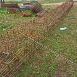 Gotowa klatka zbrojeniowa na trawie, widoczne złączenia spawane i wiązane, w tle teren budowy z ogrodzeniem i zbiornikami.