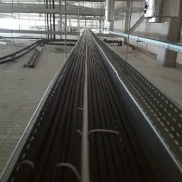 Wnętrze hali przemysłowej w trakcie montażu instalacji kablowej. Widok z góry na koryto kablowe wypełnione czarnymi przewodami, zabezpieczonymi białymi opaskami. W tle widoczne rury wentylacyjne...