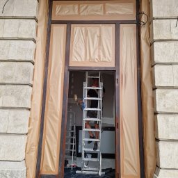 Renowacja drewna Adam Bednarski - Renowacja drewnianych drzwi wejściowych w kamienicy, zabezpieczonych papierem maskującym, malarz na drabinie z pistoletem natryskowym.