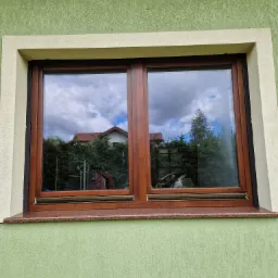 Dwuskrzydłowe okno z brązową ramą, osadzone w elewacji o zielonym odcieniu, z widocznym parapetem z kamienia w kolorze brązowo-szarym, odbijające w szybach fragment domu i drzewa