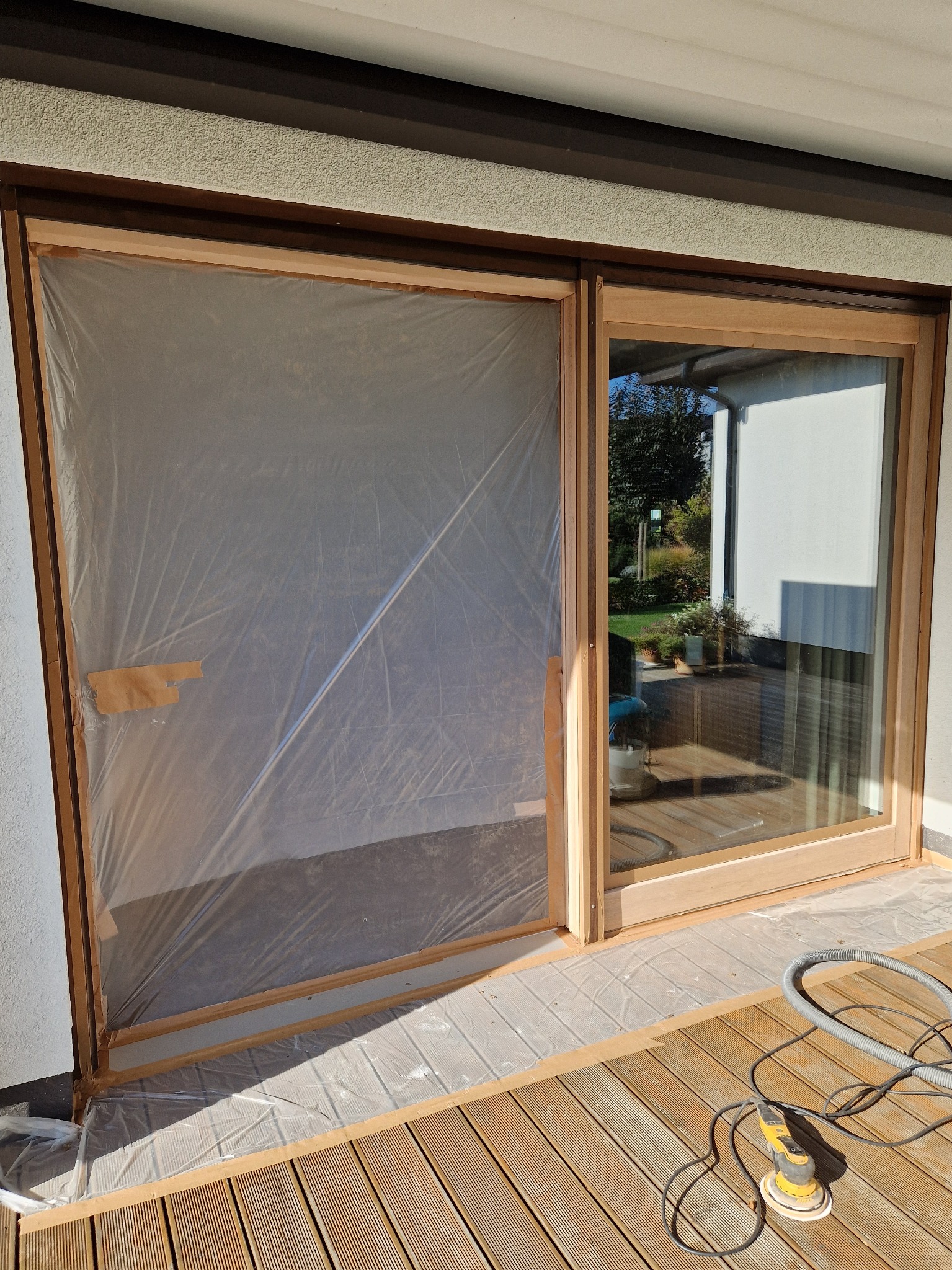 Dwu-skrzydłowe przesuwne drzwi tarasowe z surowego drewna, jedno skrzydło zabezpieczone folią malarską przyklejoną taśmą, szlifierka mimośrodowa i wąż odkurzacza na drewnianym tarasie.