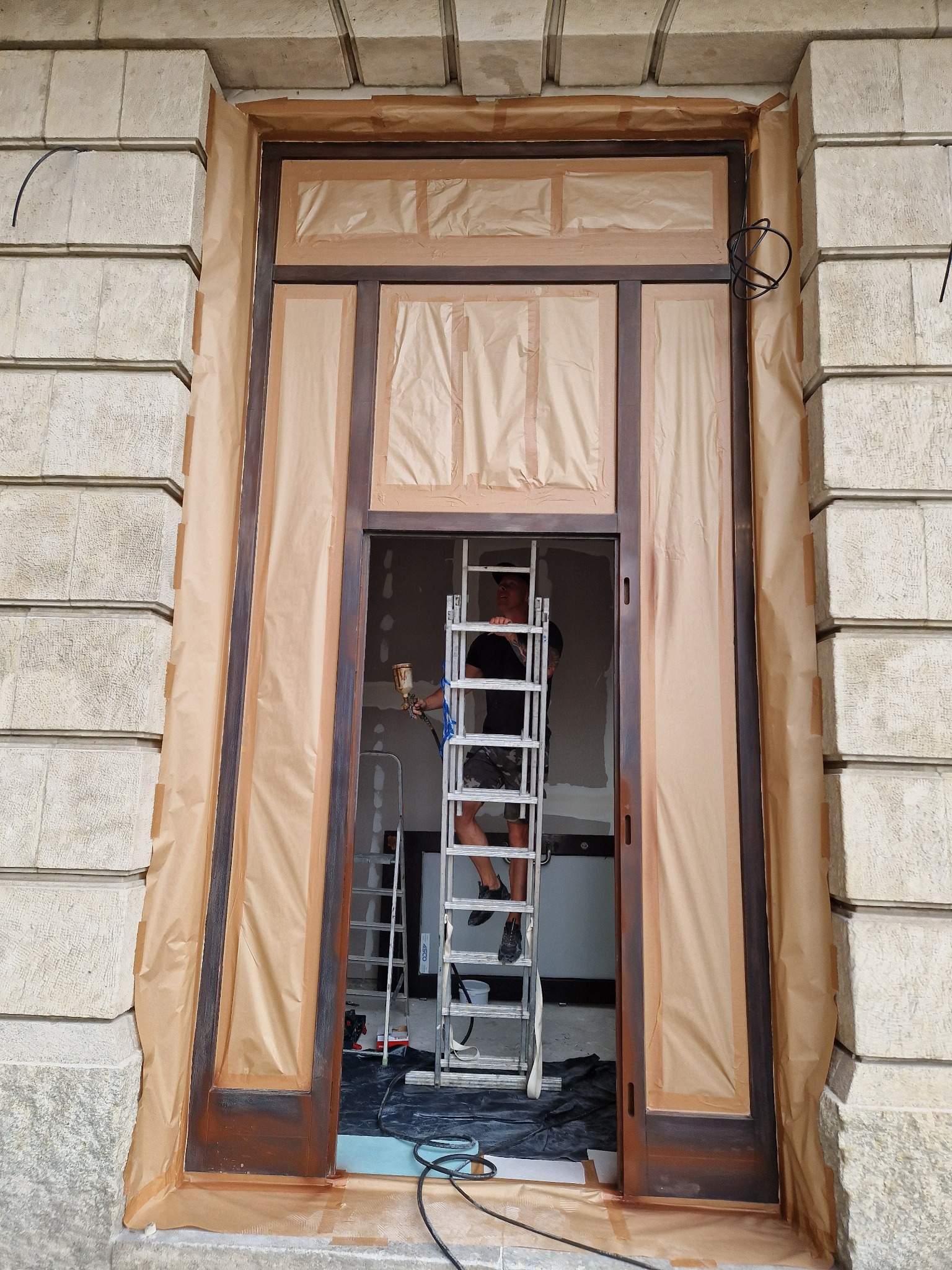 Renowacja drewnianych drzwi wejściowych w kamienicy, zabezpieczonych papierem maskującym, malarz na drabinie z pistoletem natryskowym.
