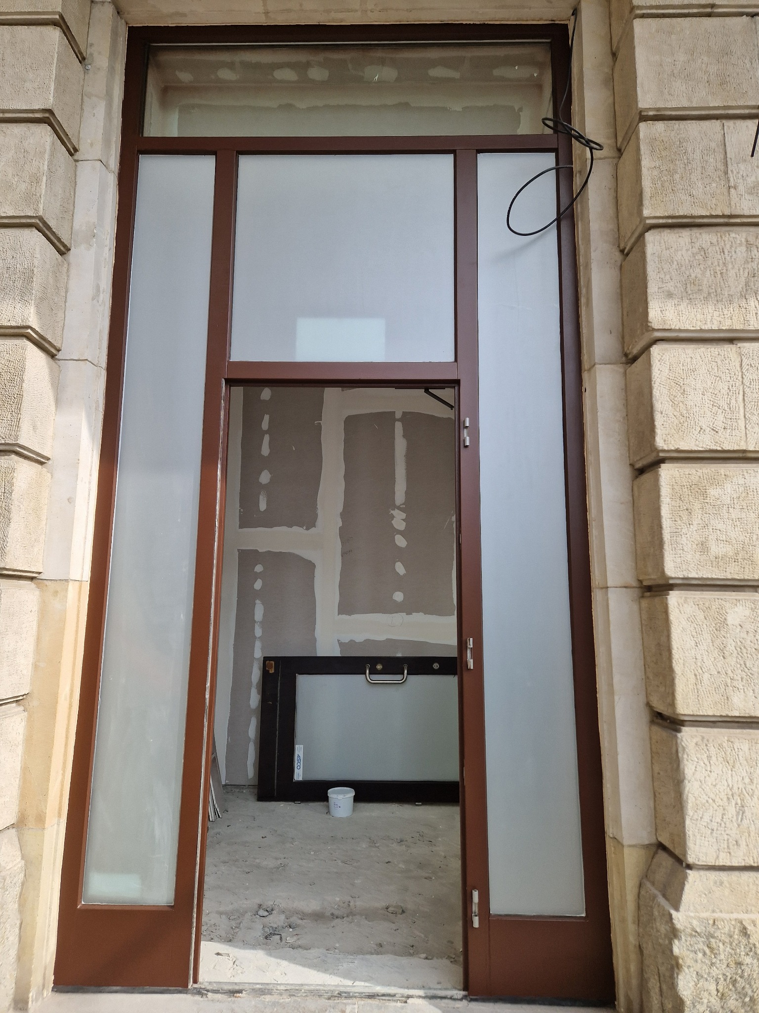 Częściowo zamontowane, brązowe drzwi z matowym szkłem, widoczne na tle surowego wnętrza z płytami gipsowo-kartonowymi i nieużywanym oknem z uchwytem.