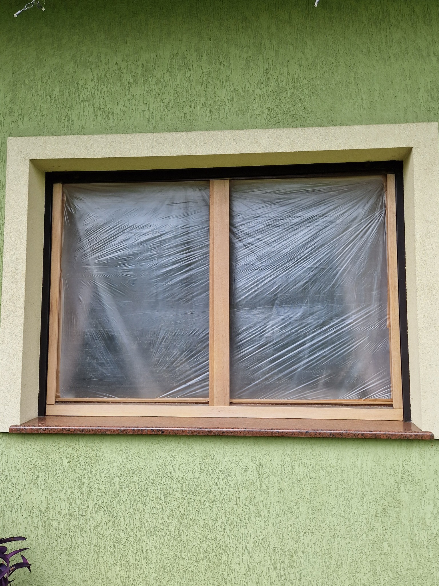 Drewniane okno zabezpieczone folią malarską podczas prac remontowych na tle zielonej elewacji.
