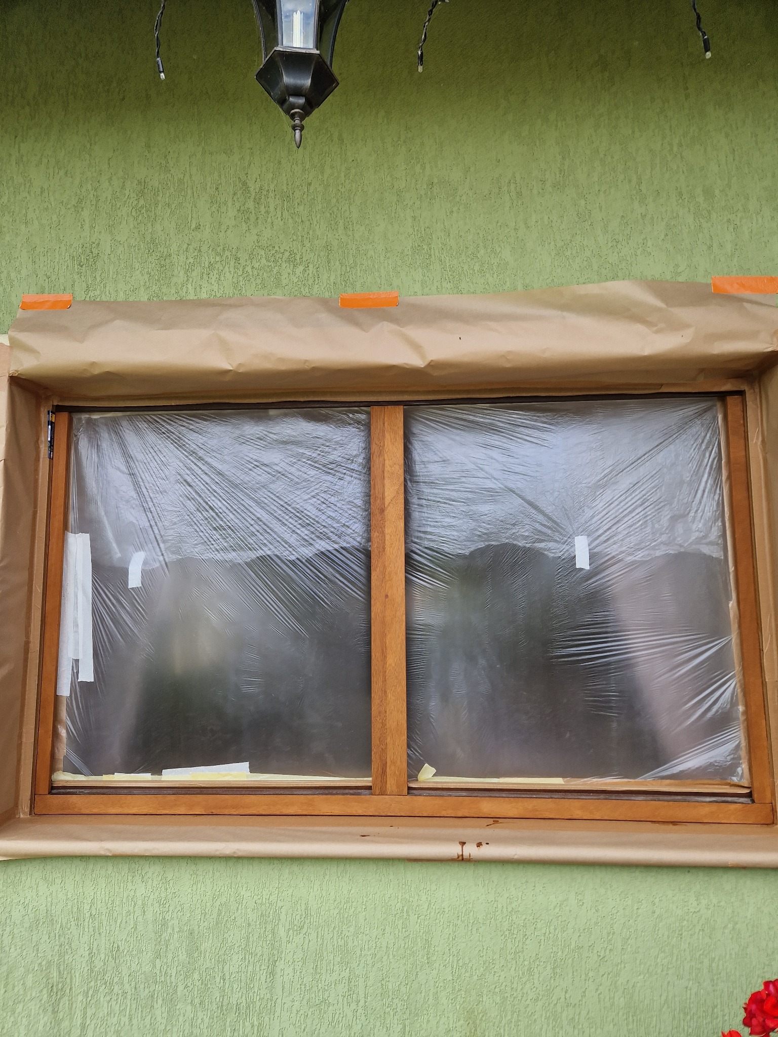 Przygotowane do malowania drewniane okno zabezpieczone folią i papierem maskującym, widoczne ślady wcześniejszych prac malarskich na elewacji. Nad oknem wisi ozdobna lampa.