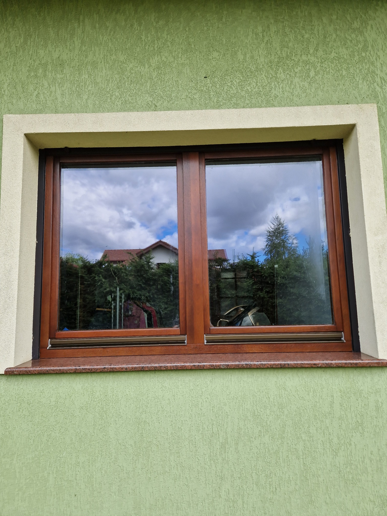 Dwuskrzydłowe okno z brązową ramą, osadzone w elewacji o zielonym odcieniu, z widocznym parapetem z kamienia w kolorze brązowo-szarym, odbijające w szybach fragment domu i drzewa