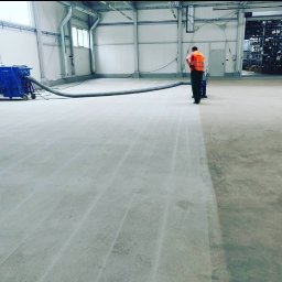 Filip Łukomski EFLOORS - Przemysłowa posadzka betonowa w trakcie czyszczenia specjalistycznym urządzeniem ssącym, pracownik w kamizelce odblaskowej, hala magazynowa w tle.