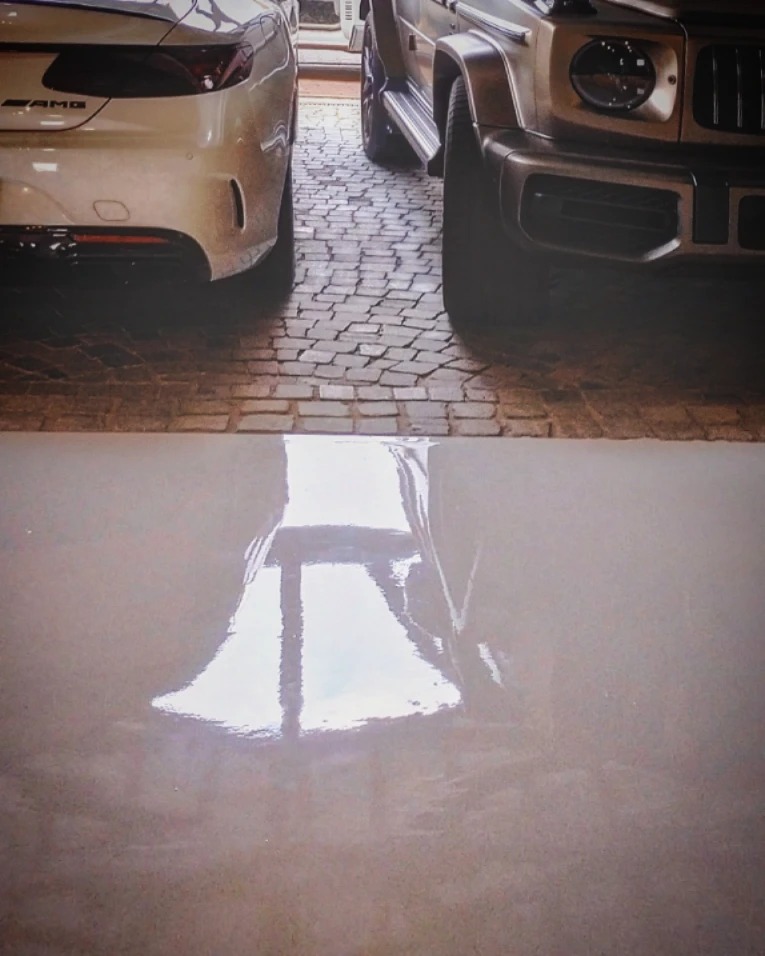 Błyszcząca, beżowa podłoga z odbiciem dwóch luksusowych samochodów; jeden biały, drugi szary, stojących na kamiennej kostce.