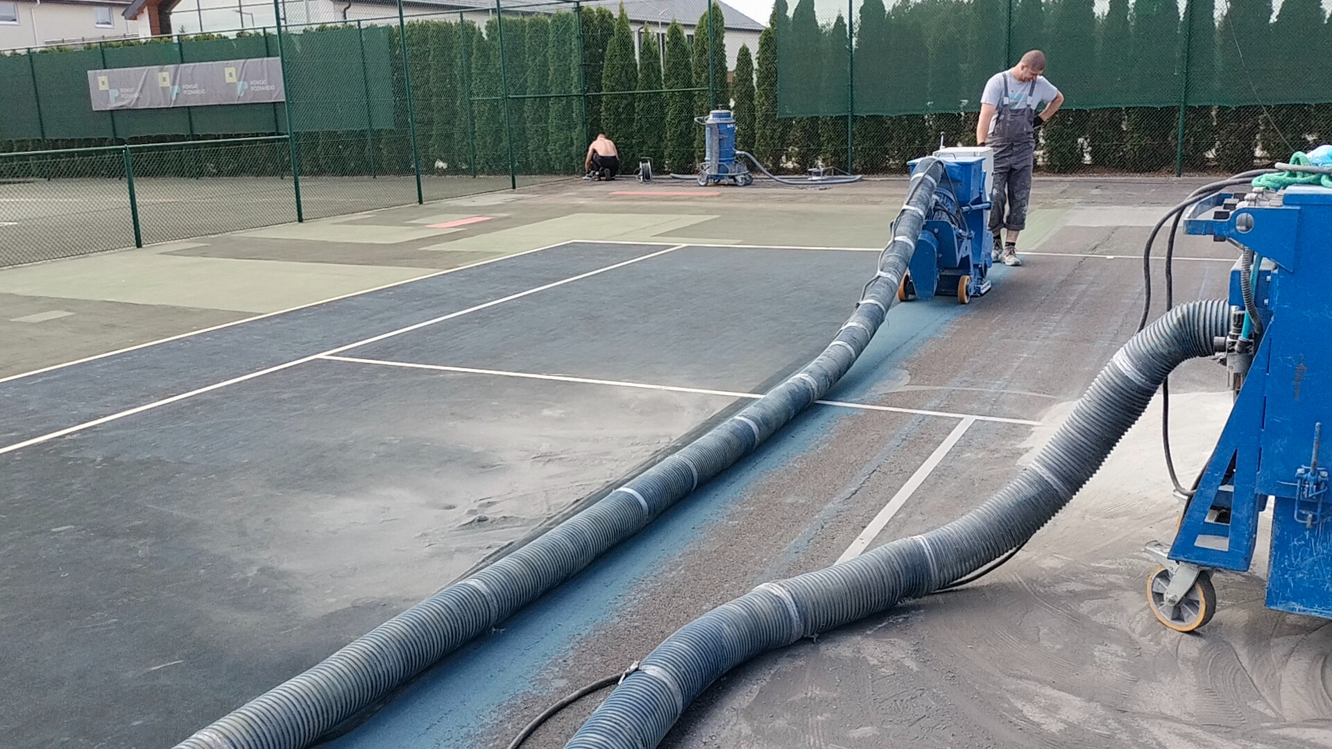 Czyszczenie nawierzchni kortu tenisowego w Kazimierzu Biskupim za pomocą specjalistycznej maszyny z systemem odsysania, widoczny pracownik w kombinezonie i drugi pracownik w tle.