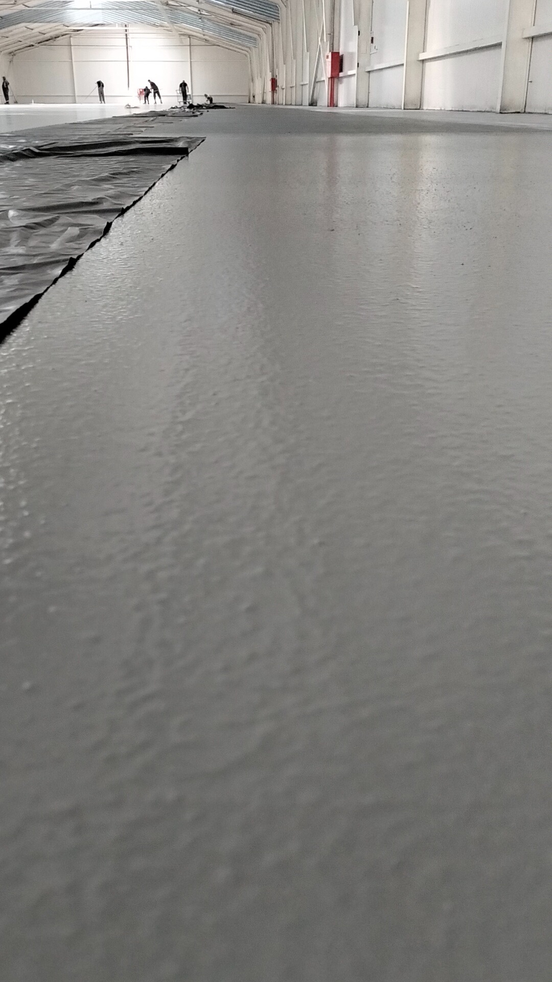 Świeżo wylana, szara posadzka w hali przemysłowej, z widoczną folią ochronną i pracownikami w oddali.