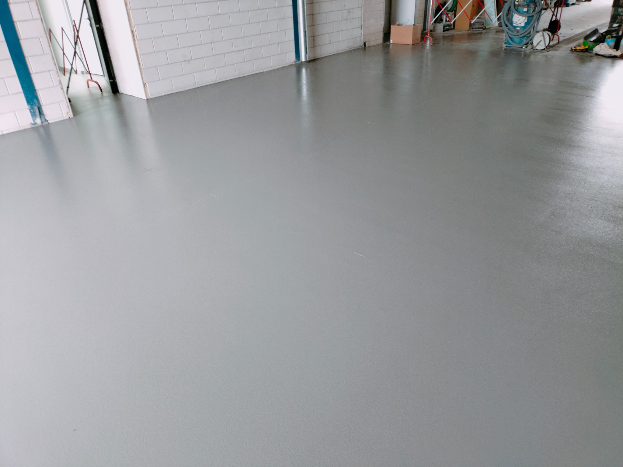 Gładka, szara posadzka w hali przemysłowej, z widocznymi akcesoriami i sprzętem w tle, przy ścianie z białych płytek.