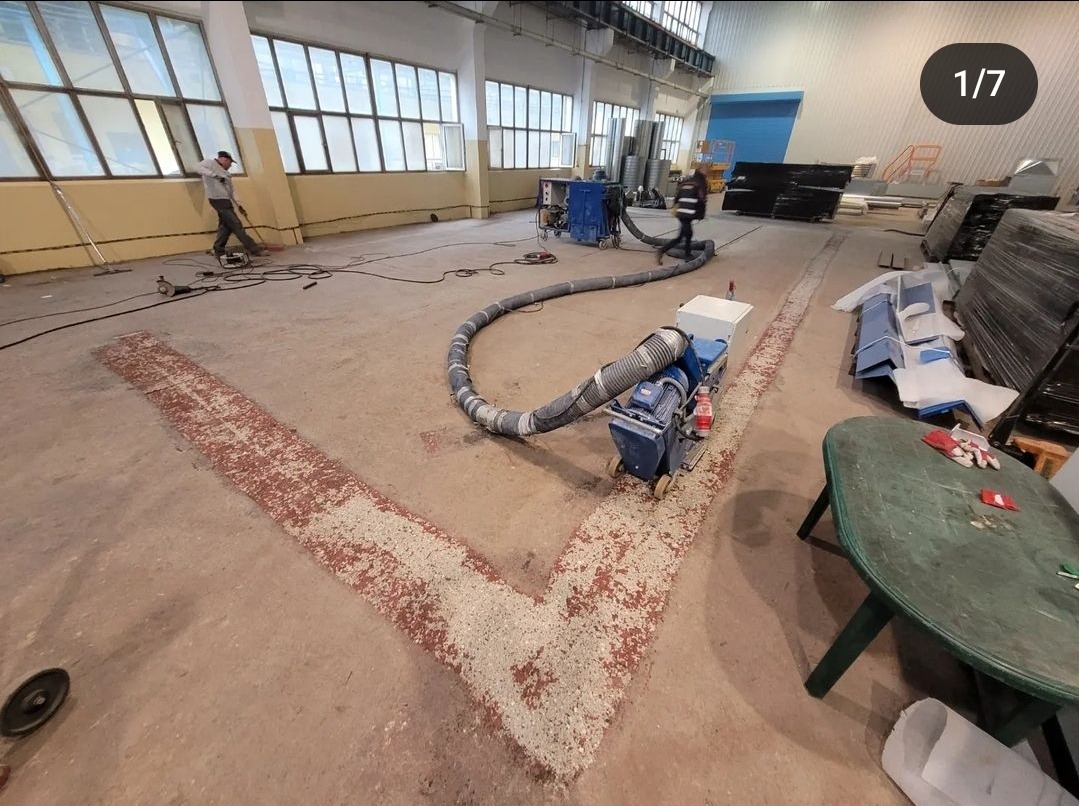 Przygotowanie podłoża w hali przemysłowej: szlifowanie posadzki za pomocą specjalistycznej maszyny z odkurzaczem przemysłowym, widoczne ślady po szlifowaniu w kształcie litery V, pracownik w tle.