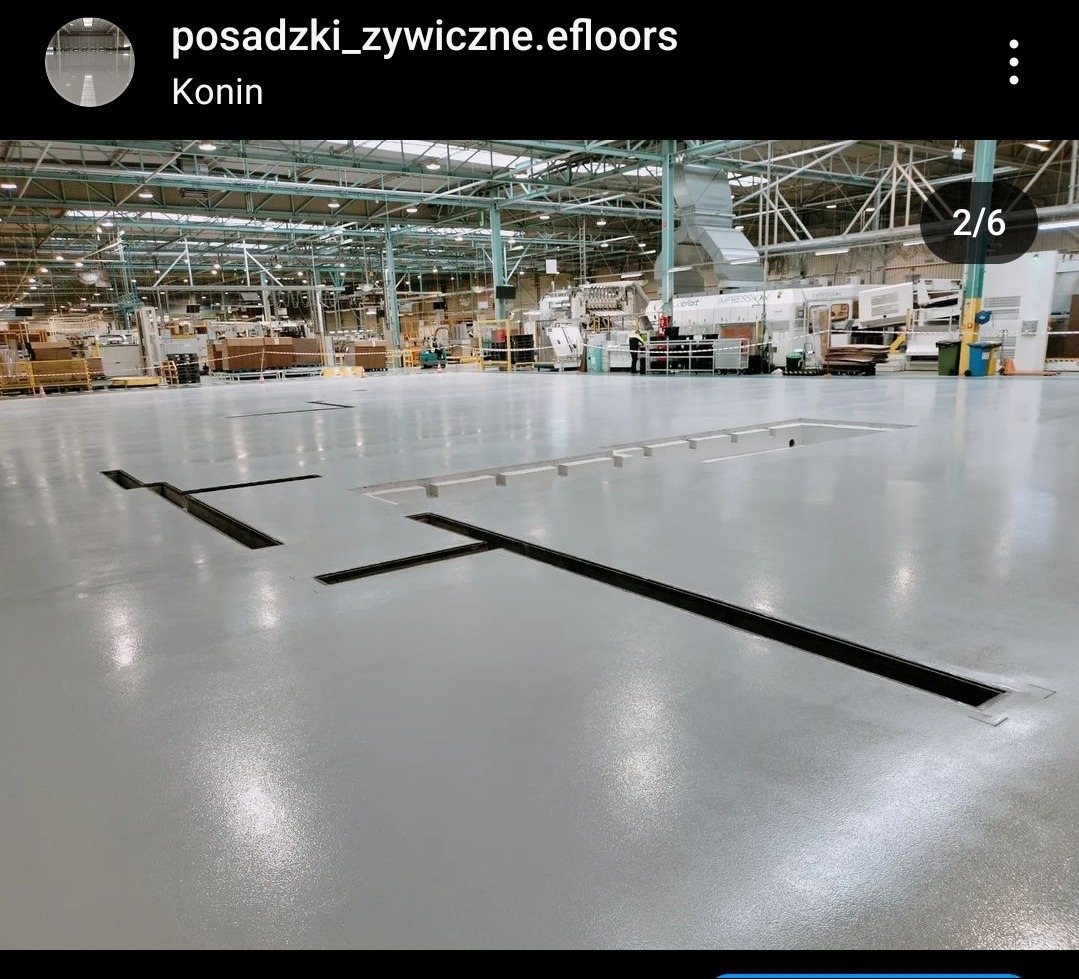 Jasnoszara, gładka podłoga w hali przemysłowej z widocznymi czarnymi szczelinami dylatacyjnymi i otworami rewizyjnymi; w tle maszyny i urządzenia produkcyjne.