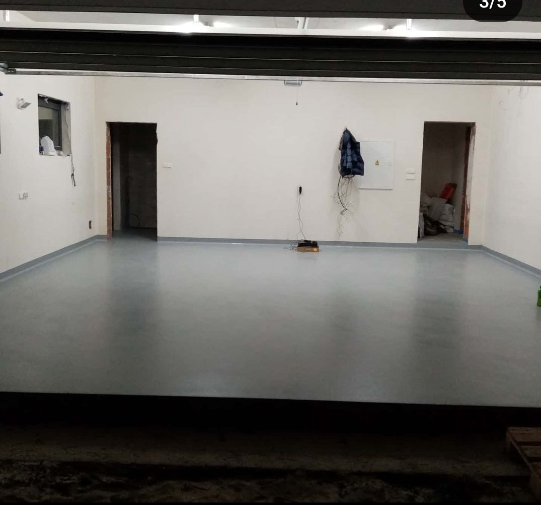 Szara, gładka posadzka w surowym wnętrzu z dwiema otwartymi przejściami, widoczna szafka elektryczna i wisząca kurtka.