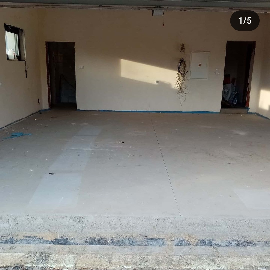 Wnętrze w trakcie prac wykończeniowych z betonową posadzką, zabezpieczonymi ścianami taśmą malarską i widoczną instalacją elektryczną.