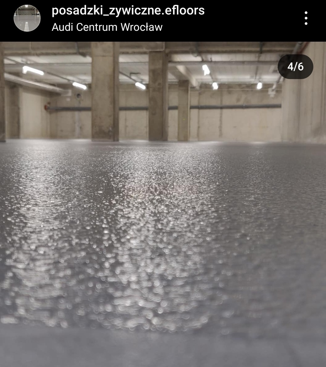 Szara, błyszcząca posadzka w surowym wnętrzu hali z betonowymi kolumnami i widocznymi instalacjami pod sufitem.