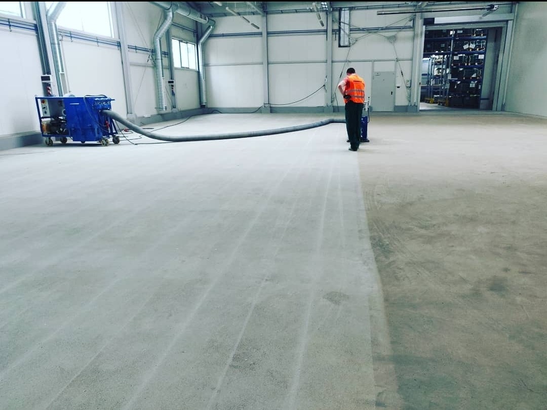 Przemysłowa posadzka betonowa w trakcie czyszczenia specjalistycznym urządzeniem ssącym, pracownik w kamizelce odblaskowej, hala magazynowa w tle.