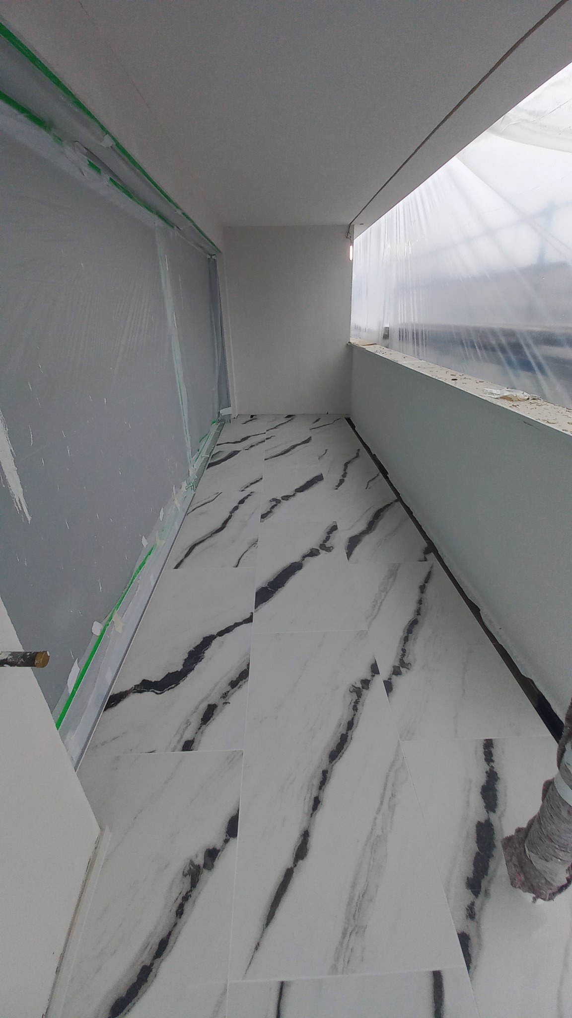 Białe płytki podłogowe z czarnym marmurkowym wzorem na balkonie w trakcie prac wykończeniowych, ściany zabezpieczone folią malarską.