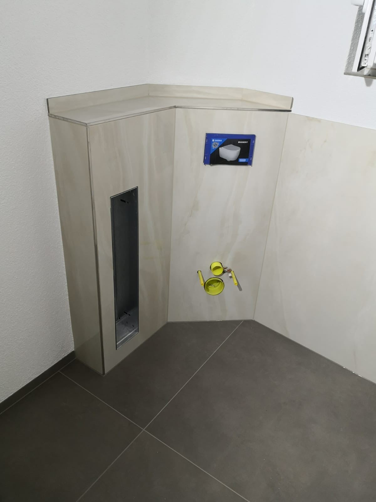 Stelaż podtynkowy WC zabudowany płytkami imitującymi marmur, widoczne przyłącza wody, otwór rewizyjny i opakowanie po misce WC.