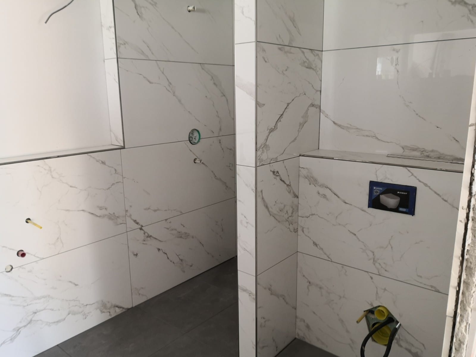 Łazienka w trakcie remontu: białe płytki imitujące marmur z szarymi żyłkami, widoczne przyłącza wodne i kanalizacyjne, szare fugi, częściowo ułożona podłoga z szarych płytek, zamontowany stelaż...