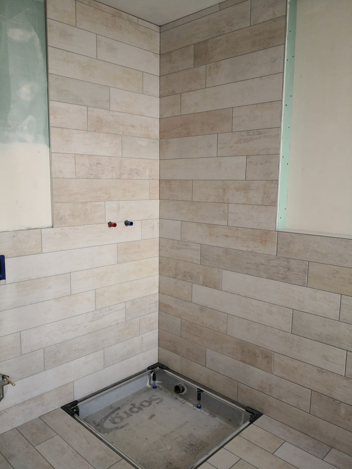 Ściany łazienki wyłożone beżowymi płytkami imitującymi drewno w układzie poziomym, z widocznym odpływem liniowym w podłodze i wystającymi rurami instalacji wodnej.