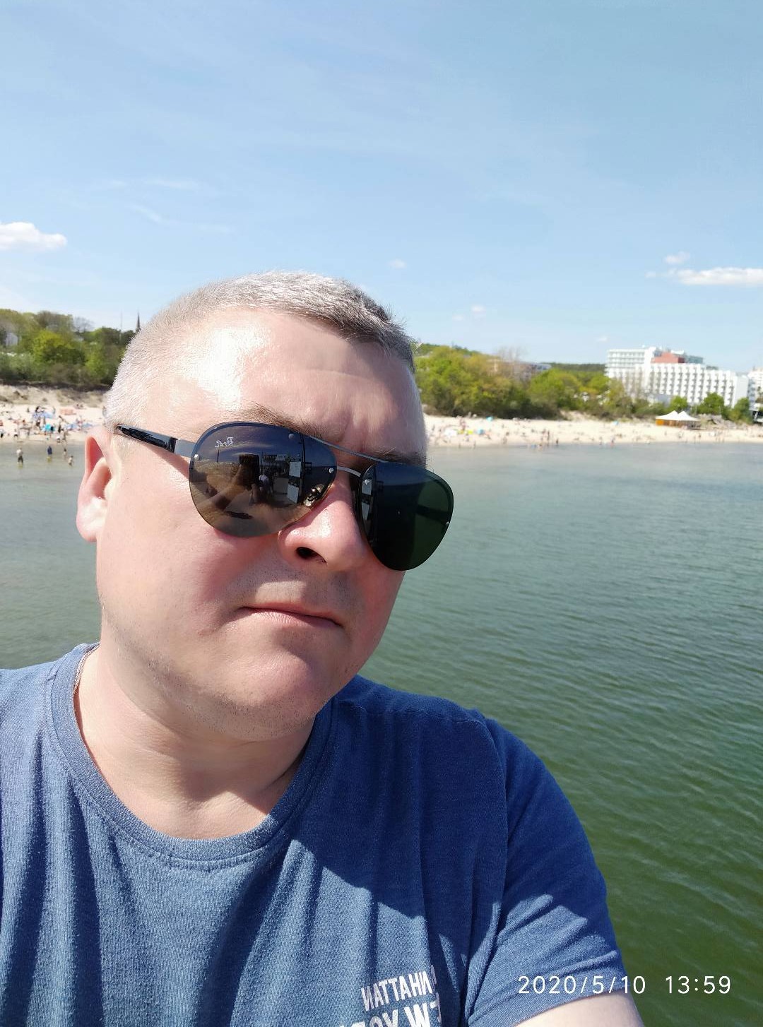 Mężczyzna w okularach przeciwsłonecznych z odbiciem plaży i budynku w szkle, na tle morza i plaży.