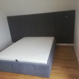 Tapicerowane łóżko z szarym wezgłowiem panelowym w pokoju ze skosami i drewnianą podłogą.