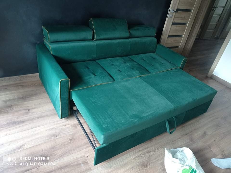 Rozkładana sofa w odcieniach zieleni z żółtą lamówką, częściowo wysunięta, na drewnianej podłodze, z widoczną ciemną ścianą i drzwiami w tle.