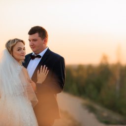 Smile! Diana Kosakowska fotografia - Para młoda w objęciach na tle zachodzącego słońca, ona w białej sukni z welonem, on w czarnym smokingu z bordową muszką, delikatne oświetlenie podkreśla ich bliskość.
