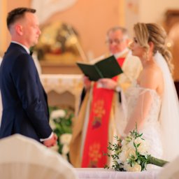 Smile! Diana Kosakowska fotografia - Para młoda podczas ceremonii ślubnej w kościele, z bukietem białych róż na pierwszym planie i księdzem czytającym z zielonej księgi w tle.