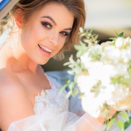 Smile! Diana Kosakowska fotografia - Uśmiechnięta panna młoda w sukni z koronkowymi detalami, trzymająca bukiet białych kwiatów, siedzi w niebieskim samochodzie i patrzy w obiektyw.