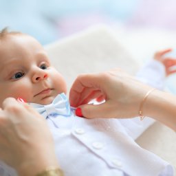 Smile! Diana Kosakowska fotografia - Niemowlę w białej koszuli i błękitnej muszce, dłoń poprawia mu muszkę, jasne tło.
