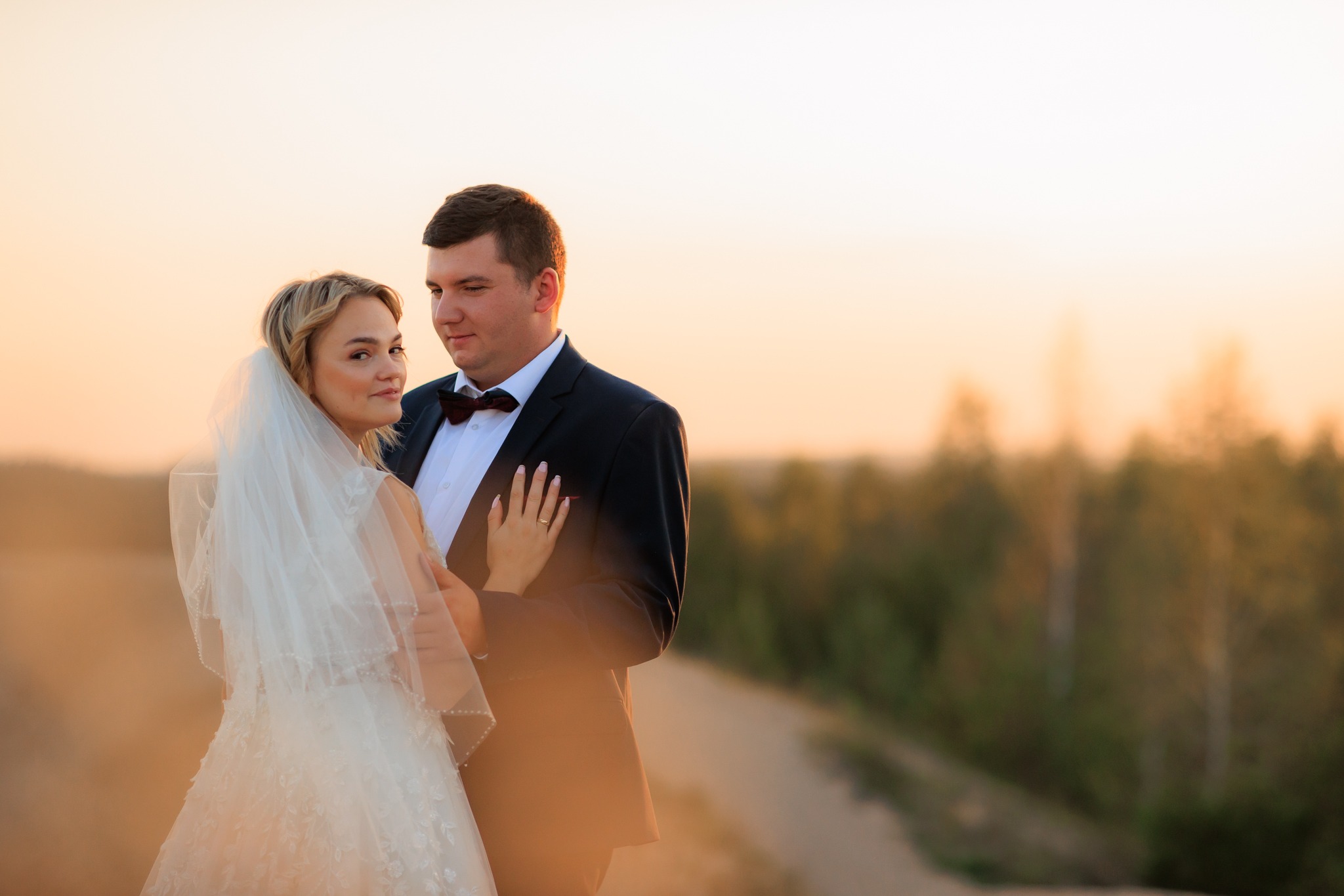 Para młoda w objęciach na tle zachodzącego słońca, ona w białej sukni z welonem, on w czarnym smokingu z bordową muszką, delikatne oświetlenie podkreśla ich bliskość.