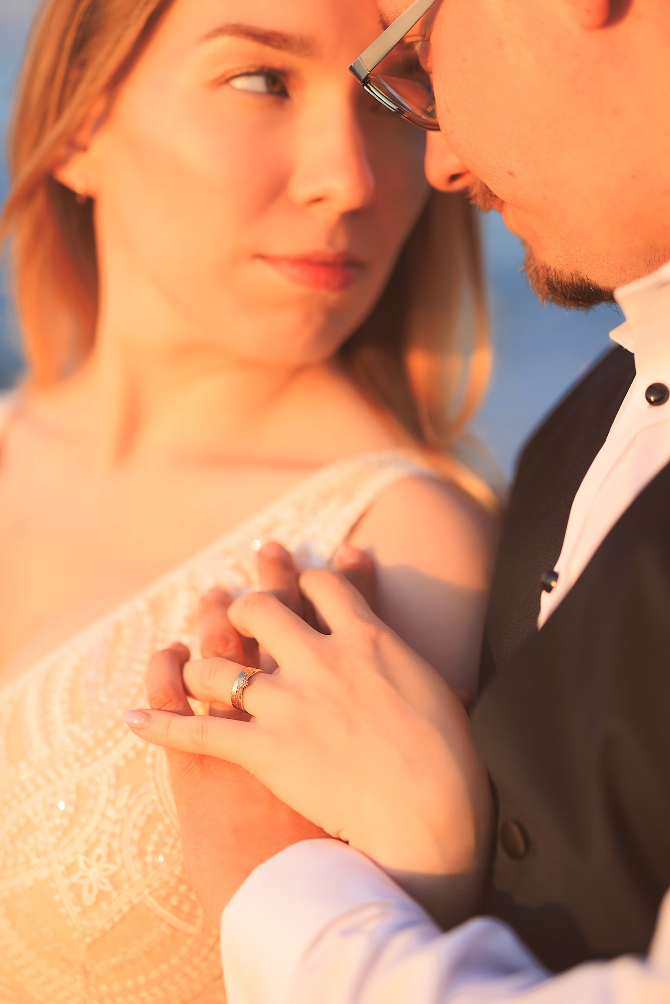 Para trzymająca się za ręce, widoczny pierścionek zaręczynowy na palcu kobiety, on w garniturze, ona w koronkowej sukni, ciepłe światło zachodzącego słońca.