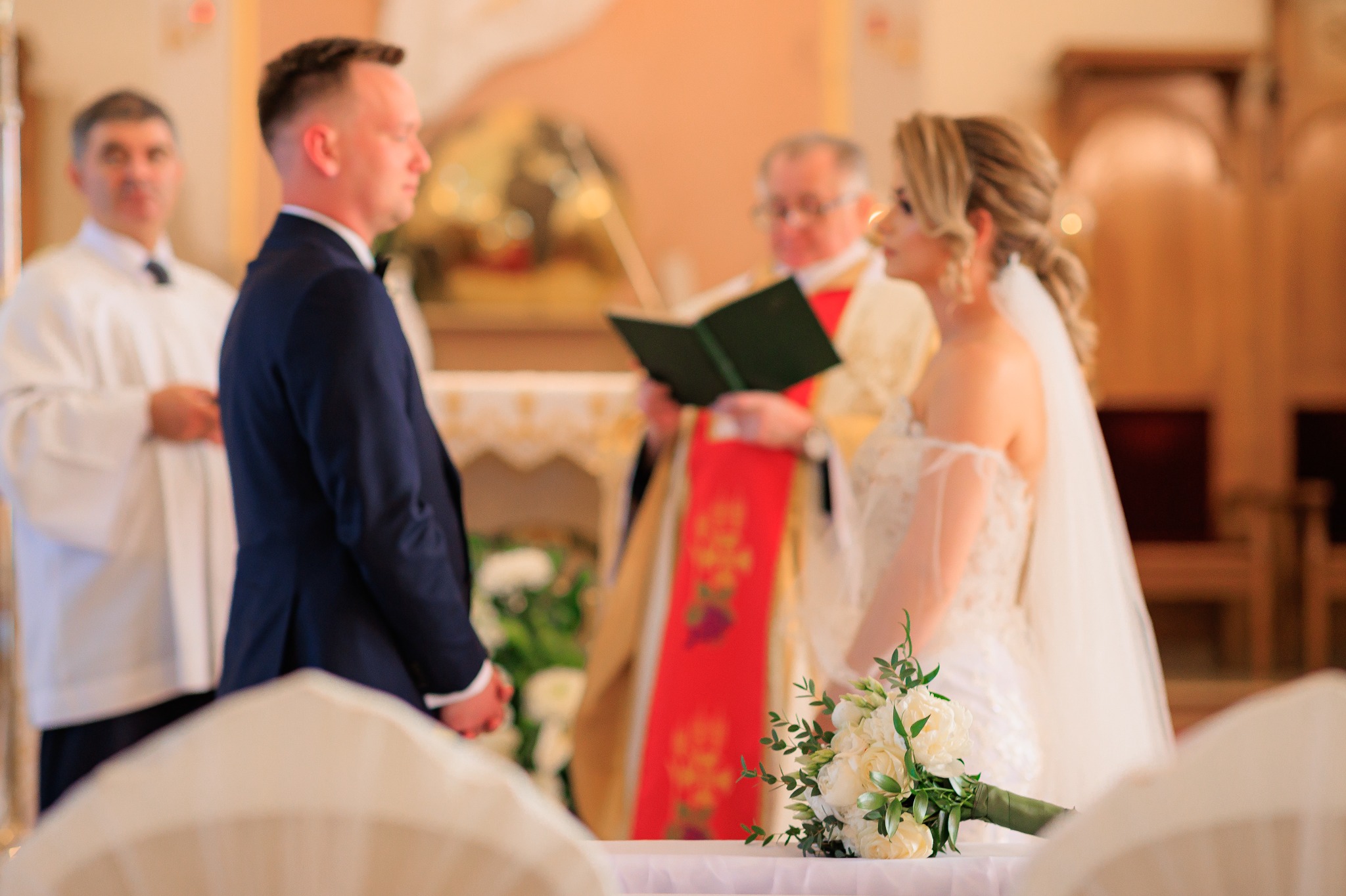 Para młoda podczas ceremonii ślubnej w kościele, z bukietem białych róż na pierwszym planie i księdzem czytającym z zielonej księgi w tle.