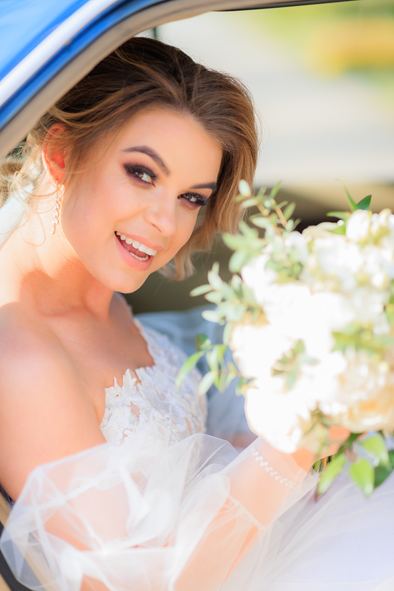 Uśmiechnięta panna młoda w sukni z koronkowymi detalami, trzymająca bukiet białych kwiatów, siedzi w niebieskim samochodzie i patrzy w obiektyw.