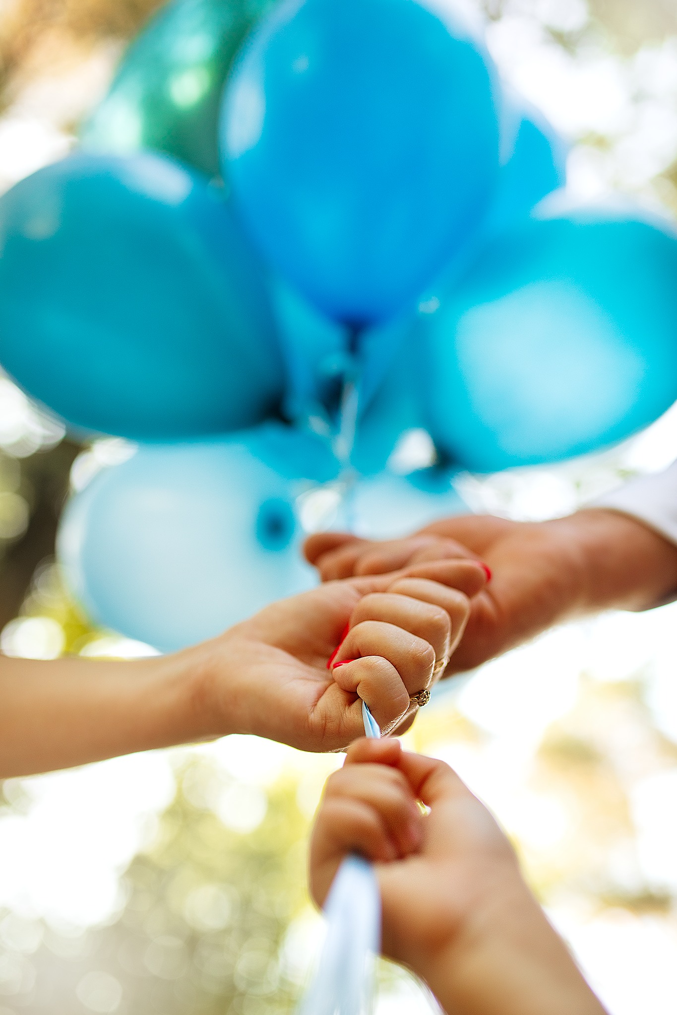 Dwie osoby trzymają wspólnie sznurek od niebieskich balonów, widoczny pierścionek na palcu kobiety, jasne tło.